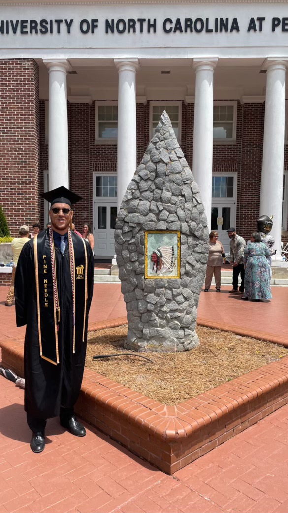 Male UNCP student in cap and gown outside of the Old Main building