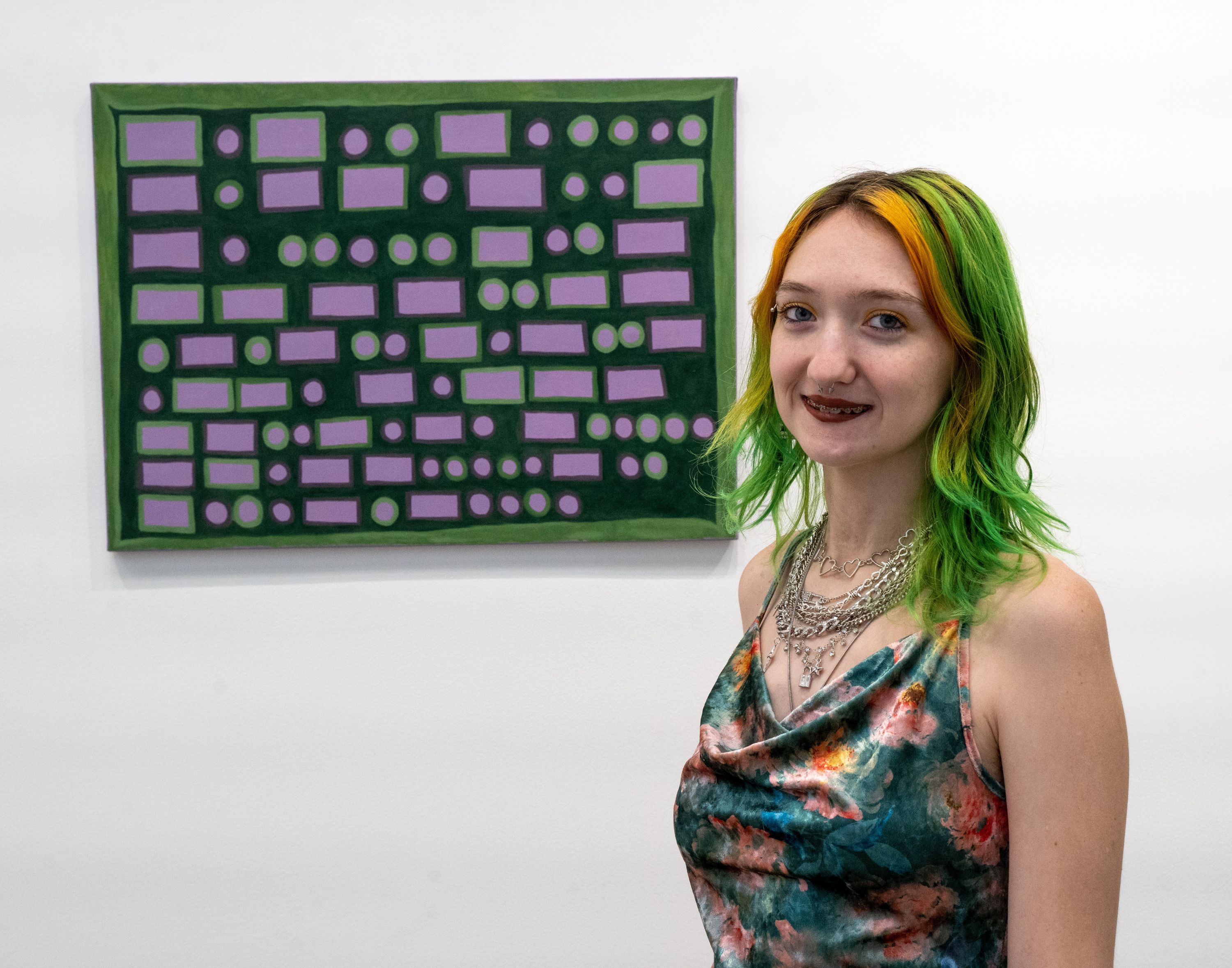 Art student, Bethani Paul, at the reception for the senior art exhibition, Hidden Lives. She's wearing a colorful dress, posing for the camera in front of her painting of morse code.