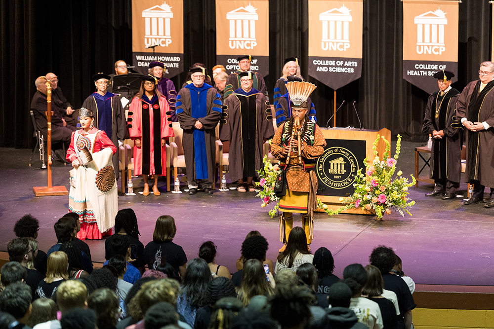 A Lumbee flutist and a Lumbee Ambassador stand on stage at Convocation