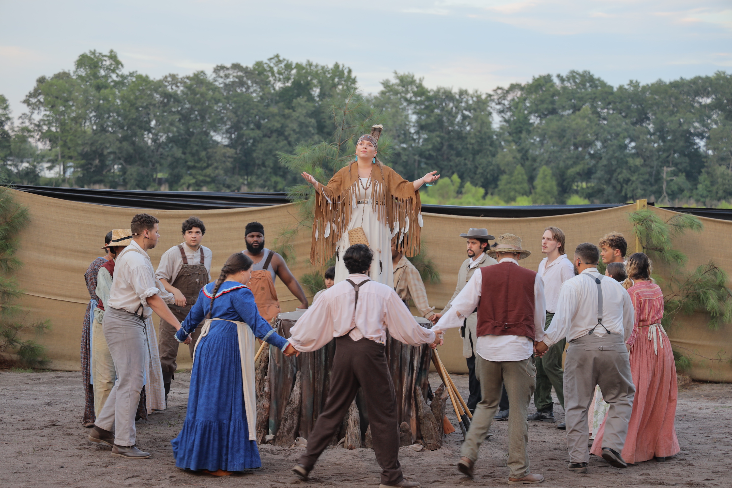 Actors stand in a circle around a woman dressed in traditional American Indian attire.