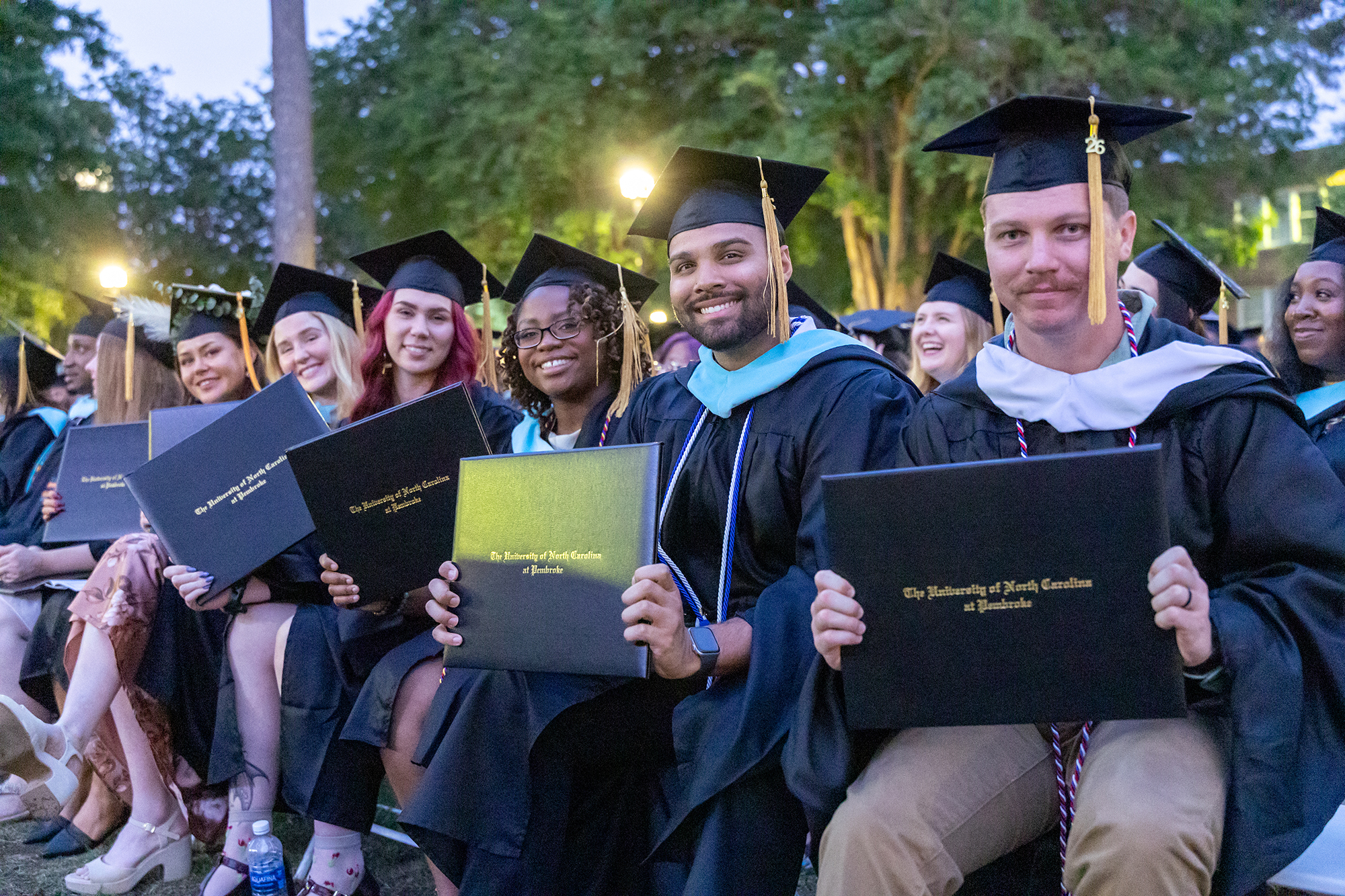 UNC Pembroke graduate students sit together outdoors during commencement, smiling and holding their diploma covers.