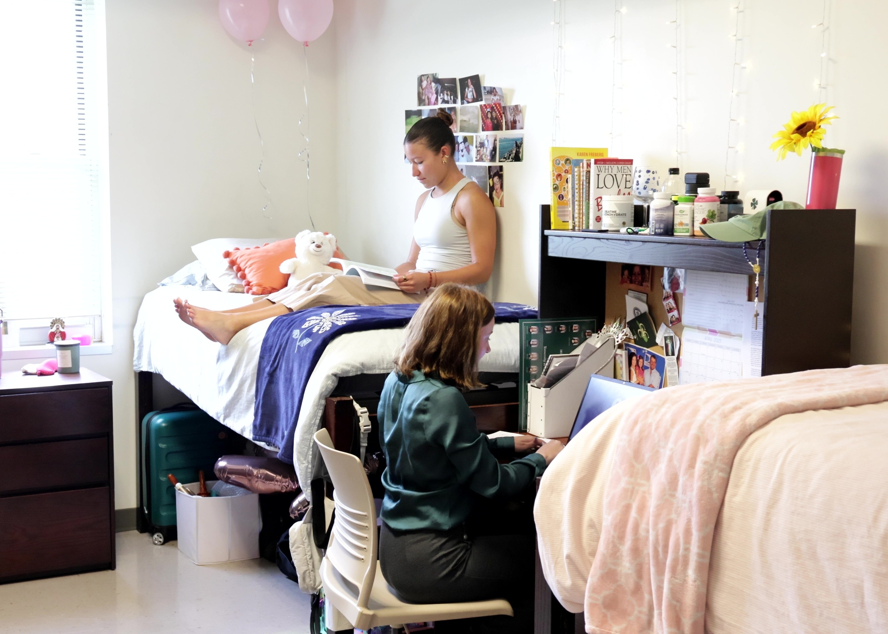 Two female students sharing a dorm room, Cypress Hall at UNC Pembroke. One sits up reading on a high bed. The other is seated at a desk with a laptop.