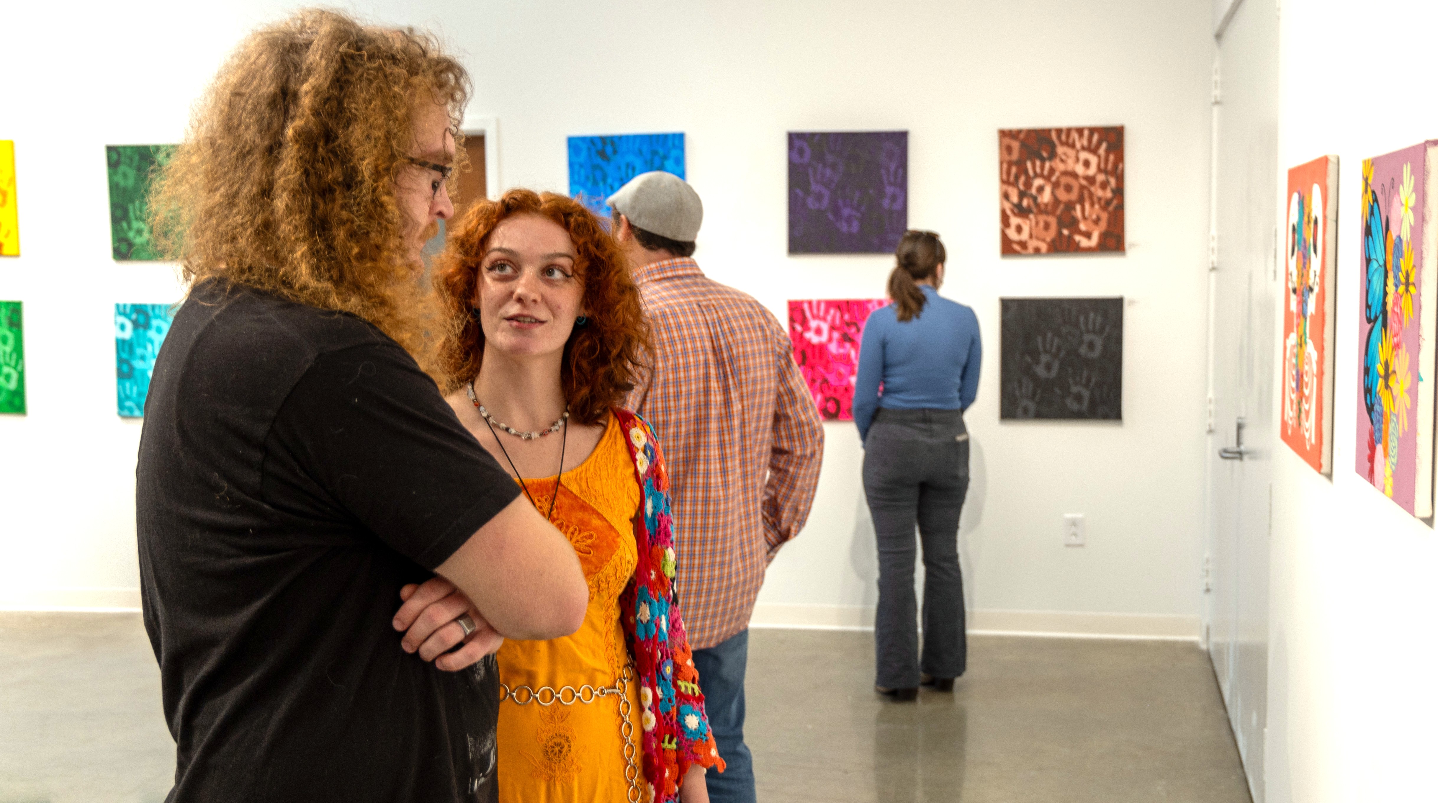 Two college students talk while admiring paintings mounted on the wall in front of them. Other students and public stand in the background looking at other works on other walls.