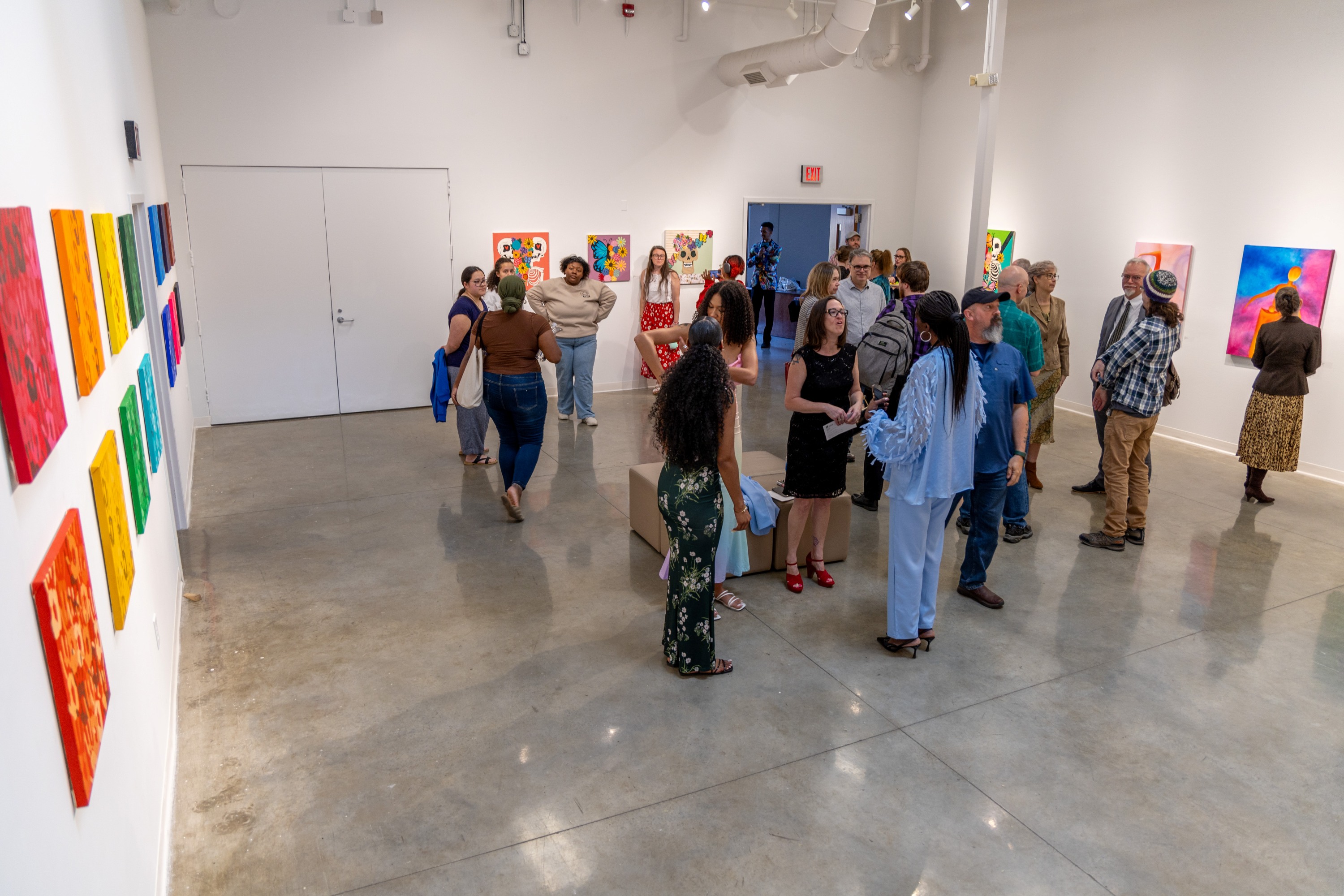Art gallery reception full of people talking and looking at art mounted on white walls. Colorful paintings line the left wall shown in the image.