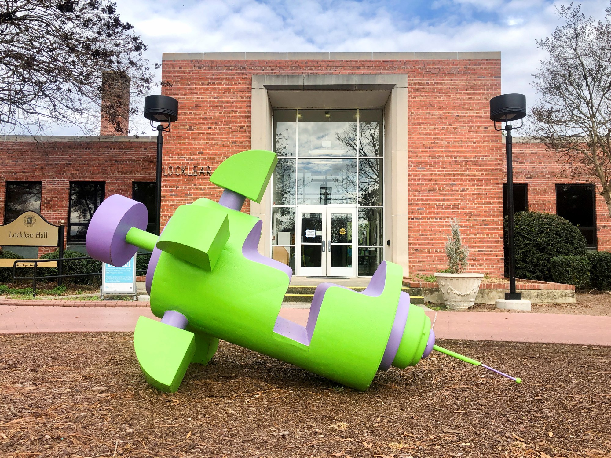 A large sculpture of a cartoonish syringe with seating for two as if it were a ride. The sculpture rests in front of Locklear Hall, home to the A.D. Gallery.