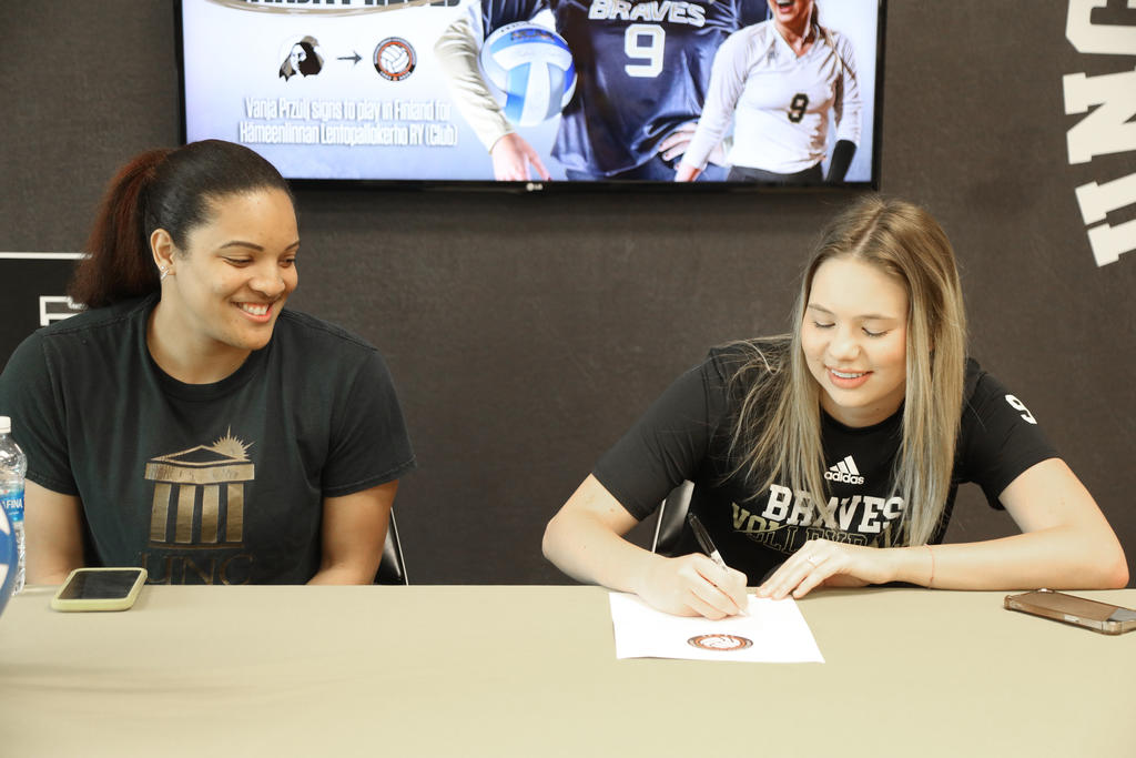 UNC Pembroke volleyball player signs professional contract while seated beside a smiling coach, with a screen in the background announcing the signing.