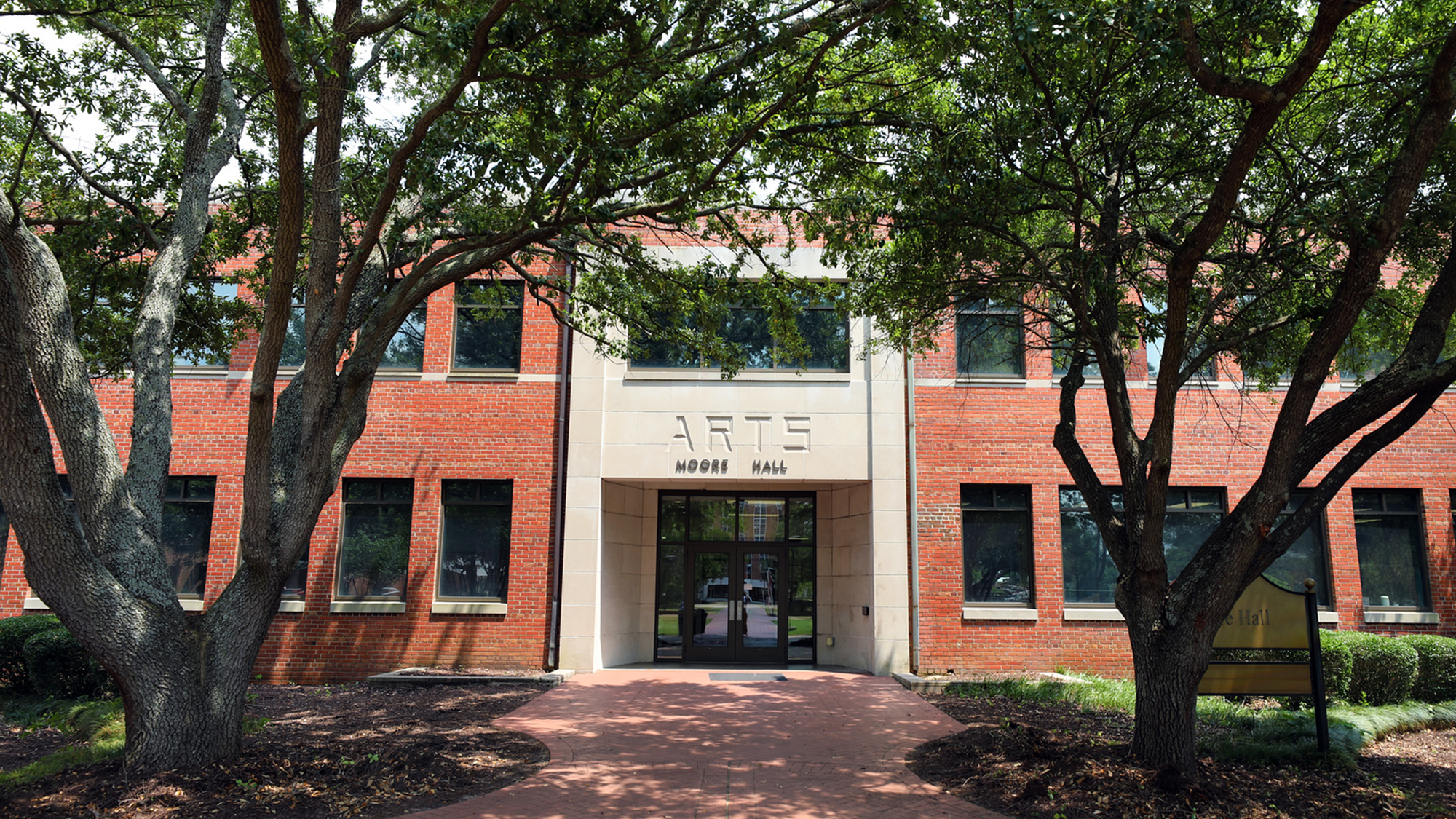 Front entrance of Moore Hall, home to the Department of Music and Arts at UNC Pembroke, framed by large trees and a brick walkway on a sunny day.