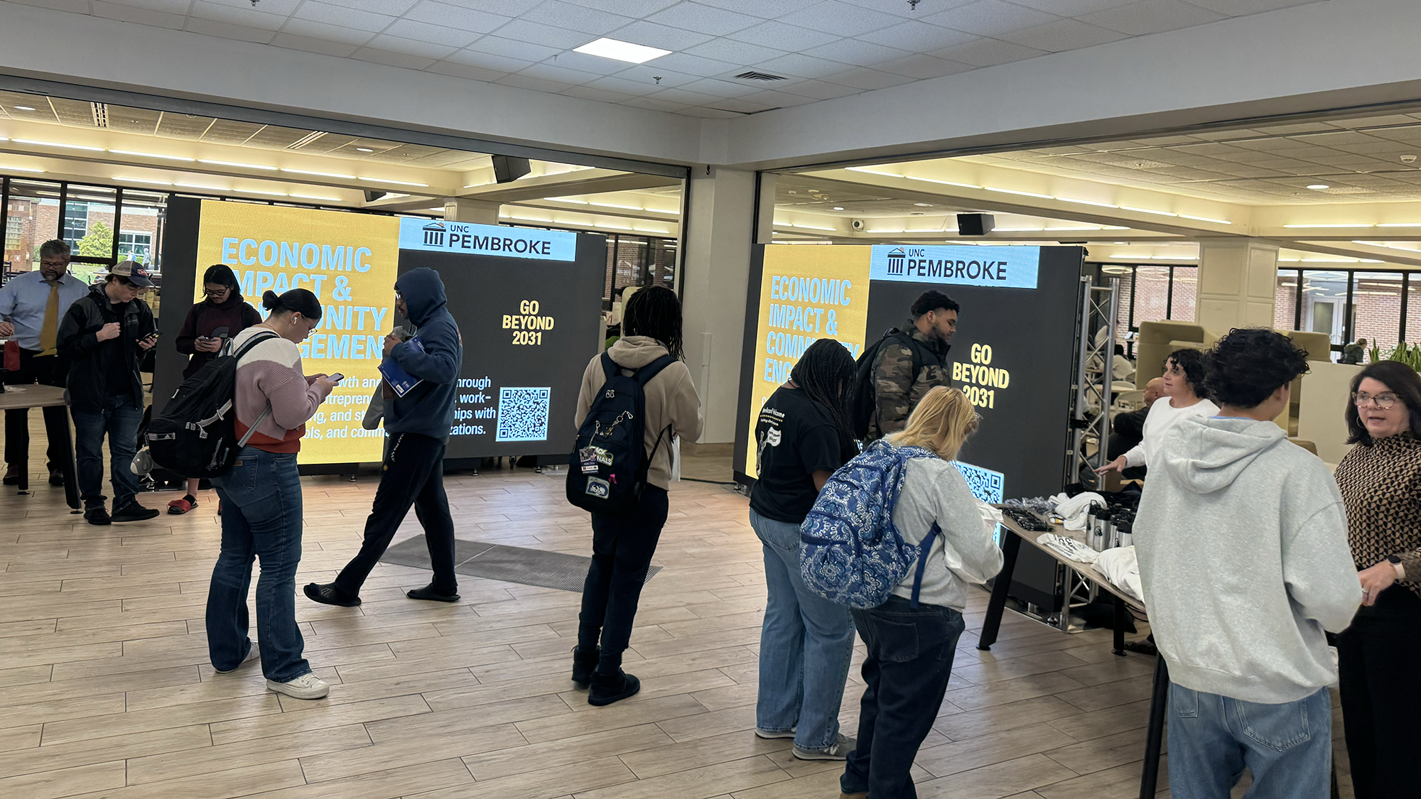 Students,faculty and staff stand in a commons area near large screens participating in a charrette.