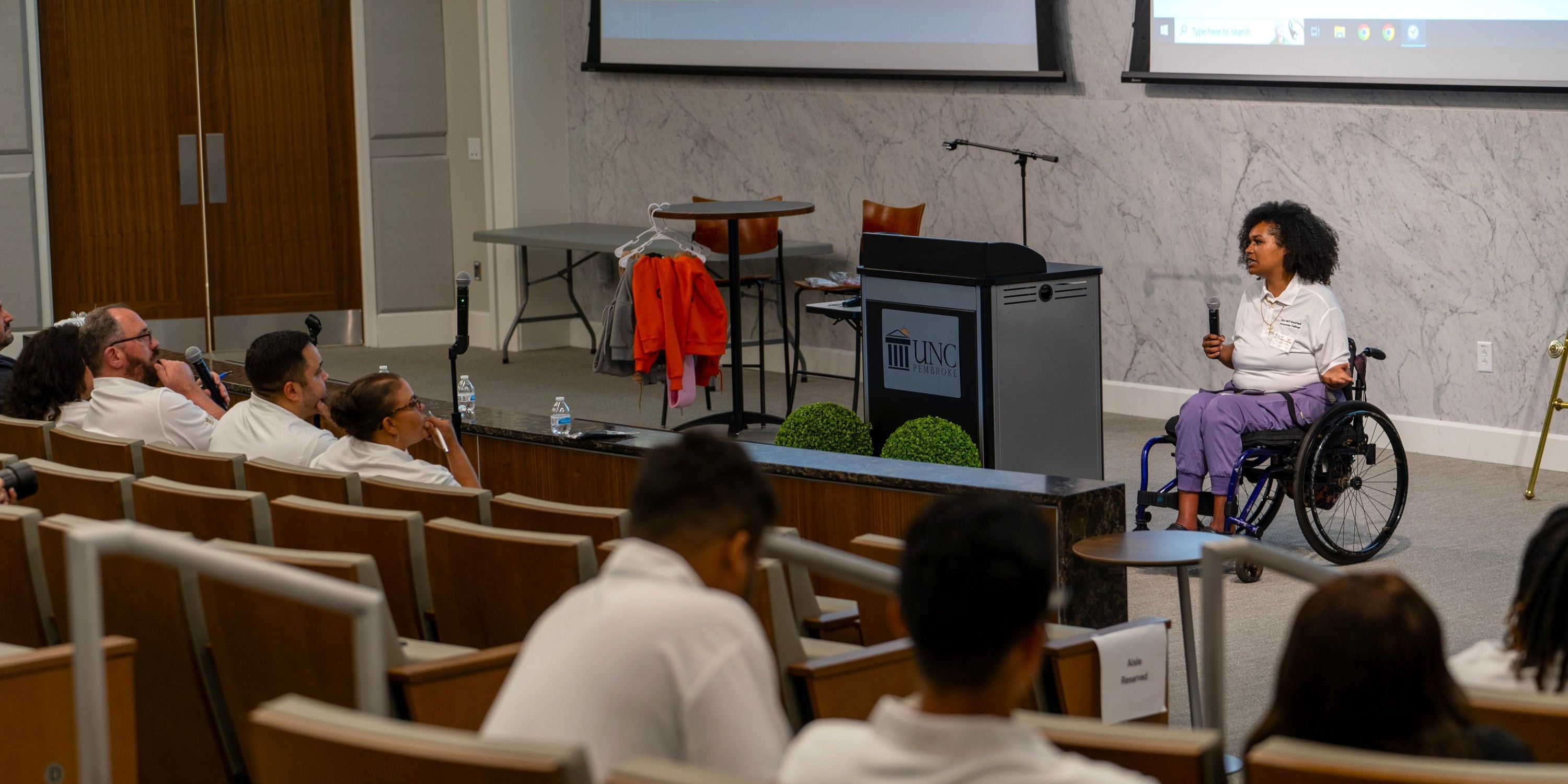 Female student in a wheelchair presenting her entrepreneurship pitch to a panel of judges and fellow students. Her product pitch is for an innovative whelchair accssory that provides rain cover for persons in wheelchairs. The event was the BraveHawk Entrepeneurship Challenge, by the Thomas College of Bursiness and Economics at UNC Pembroke.