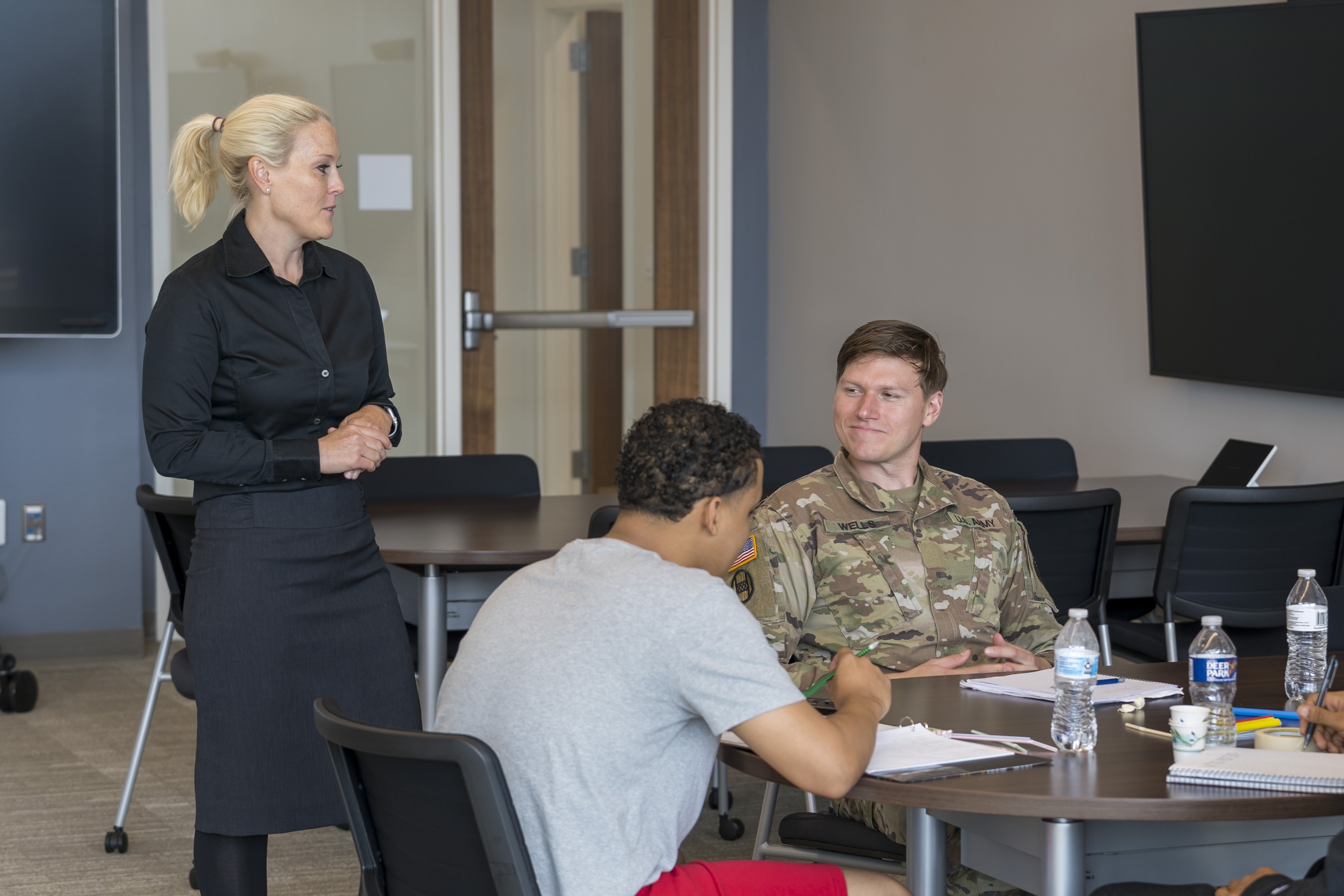 Classroom image of a female professor talking with three students at a table. A male ROTC student in military fatigues smiles at her during the conversation.