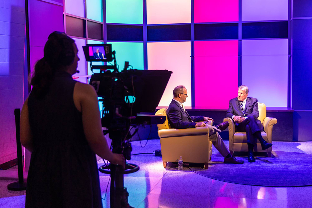 A student camera operator films two men seated in armchairs during a studio interview, with a colorful backlit set behind them