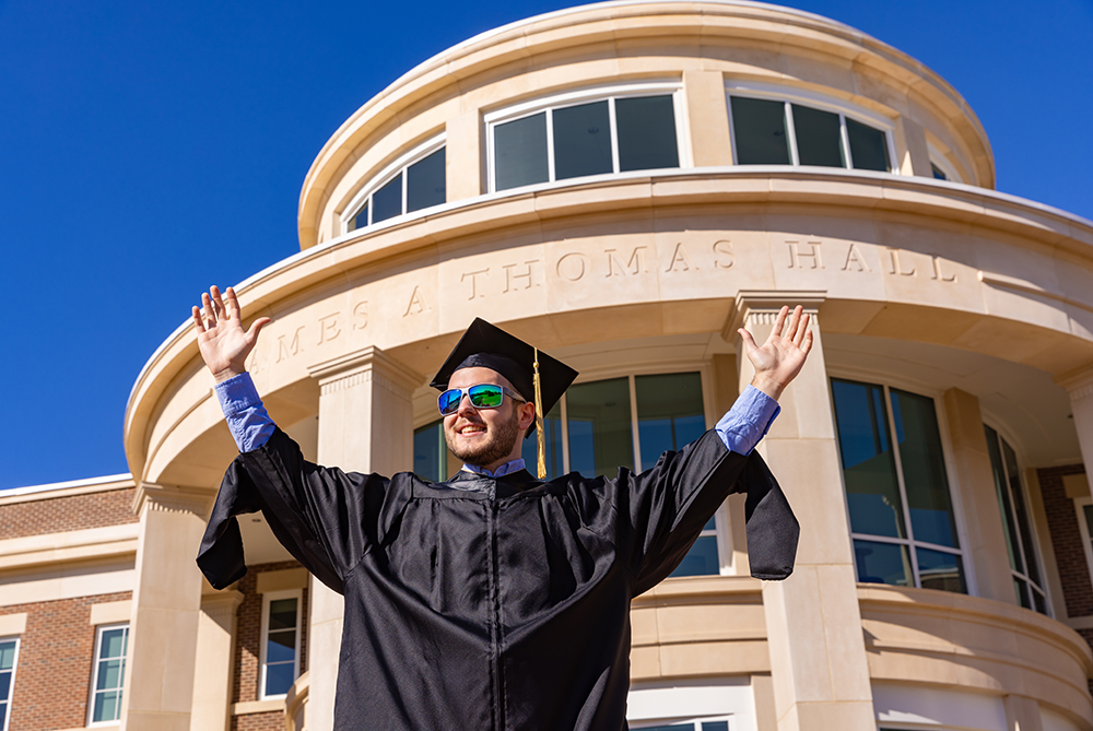 Trey male student graduation cap and gown in front of James A. Thomas Hall with arms up