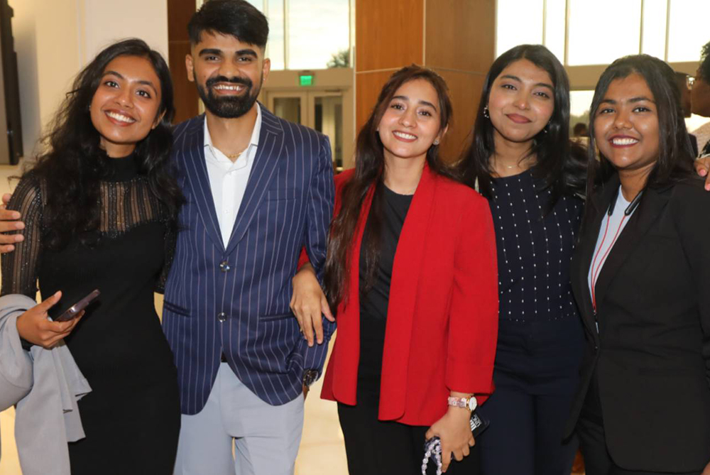 Five international students dressed up for an event, standing with each other and smiling at the camera. Two of the five have their arms around the others.