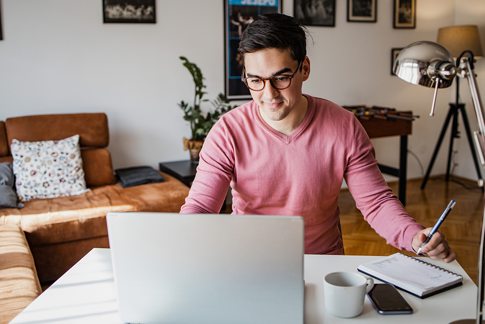 Young man wearing glasses in a long-sleeved pink v-neck shirt, in a living room space, at a table with a laptop open in front of him with a notebook at the side, a pencil in hand.