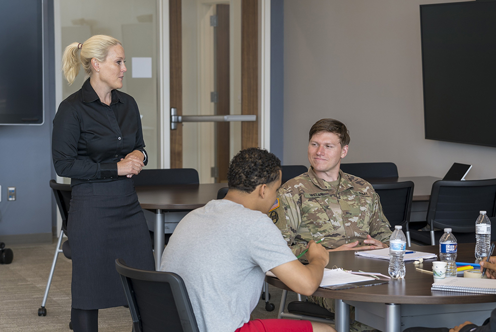 Classroom image of a female professor talking with three students at a table. A male ROTC student in military fatigues smiles at her during the conversation.