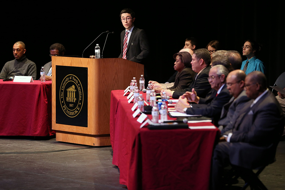 UNCP SGA president at a podium, winged by two long tables of local school board representatives, leading a debate.