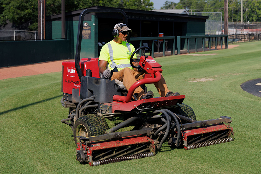 UNC Pembroke facilities and maintenance staff wearing the appropriate protective gear on a large riding mower cutting grass on the baseball field.