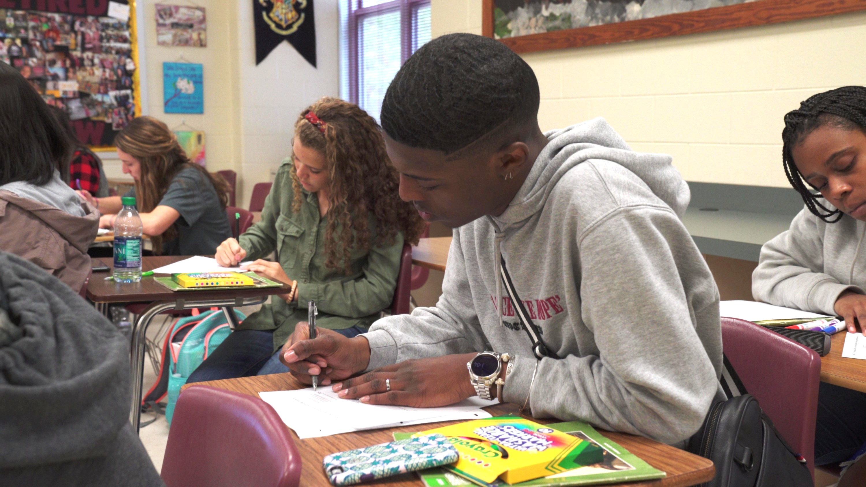 Male high school student in the classroom at his desk working on a worksheet. Female calssmates are visible at desks behind and beside him also on their worksheets.