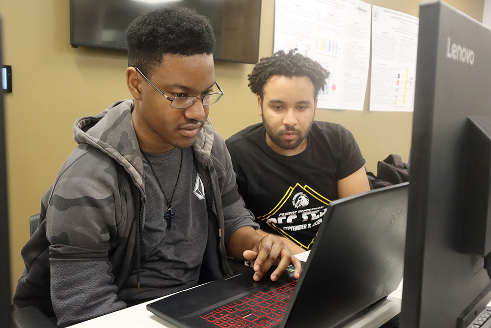 Two black male students hunched over a laptop, intently looking at the screen.