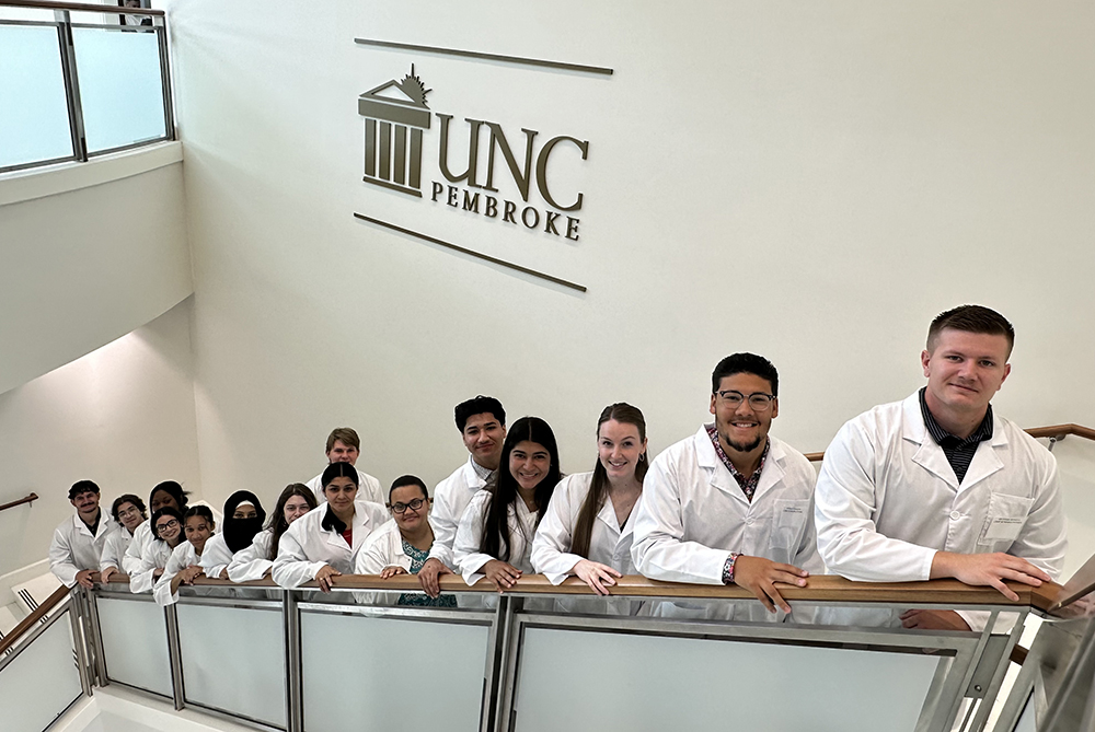 Group of medical students or interns in scrubs gathered around a table, listening to a peer giving a presentation at a whiteboard.