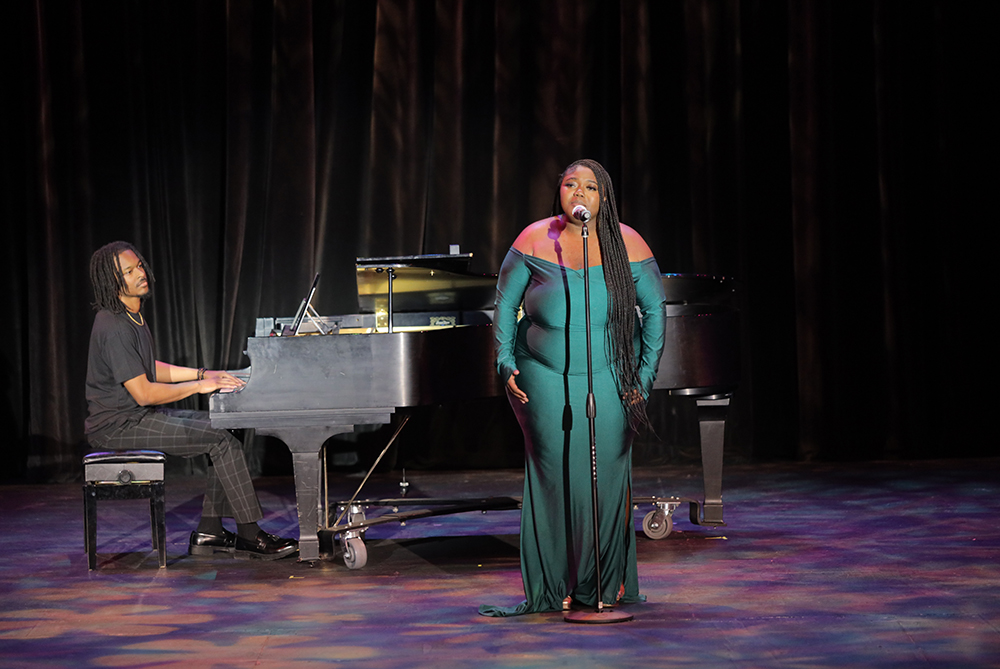 A vocalist in a green gown performs on stage with a pianist accompanying her on a grand piano during a talent show.