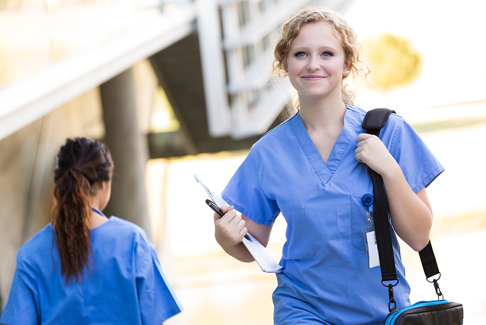 a woman in scrubs walking down through street with a folder tucked in her arm smiling
