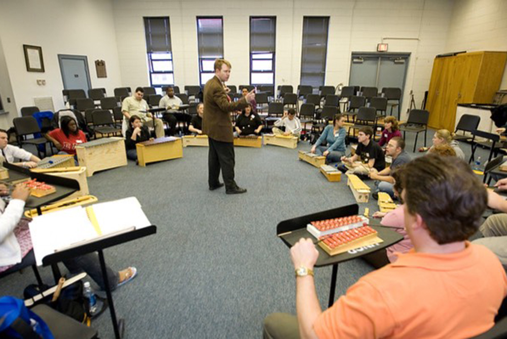 Music education students seated in a circle playing percussion instruments while an instructor leads a classroom session, demonstrating hands-on teaching methods at UNC Pembroke.