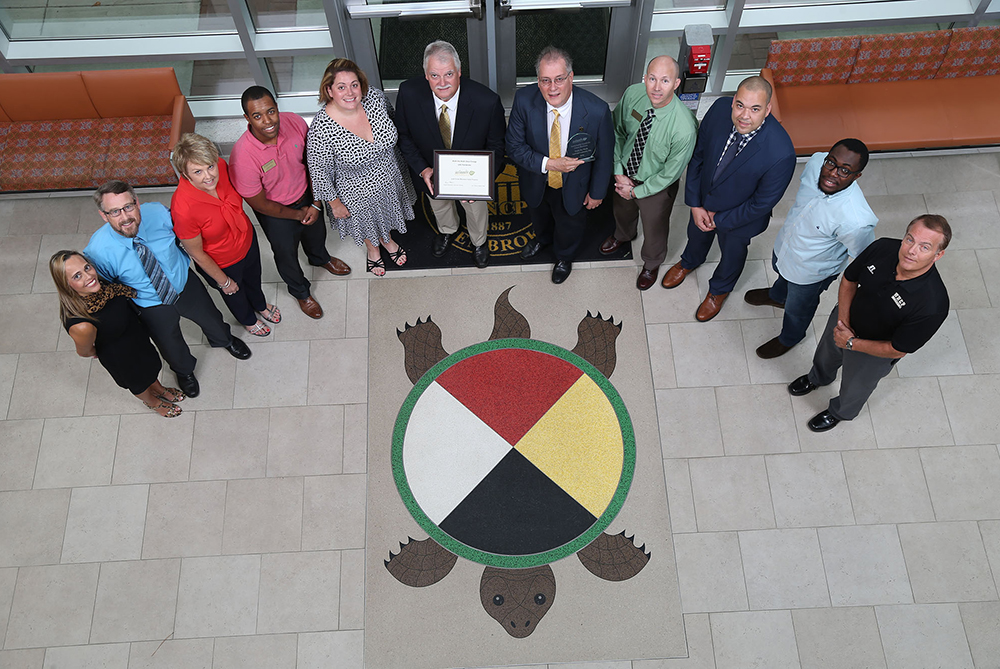 A group of eight people stands smiling in front of a “Tree Campus USA” banner on a university campus near a small pond. The individuals, including students and faculty, are gathered around a newly planted tree wrapped with a gold and black bow.