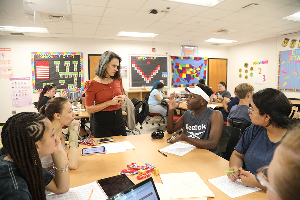 A classroom with several students seated at tables, working or discussing in groups. A teacher stands and interacts with one group. The walls are decorated with educational posters and colorful bulletin boards.