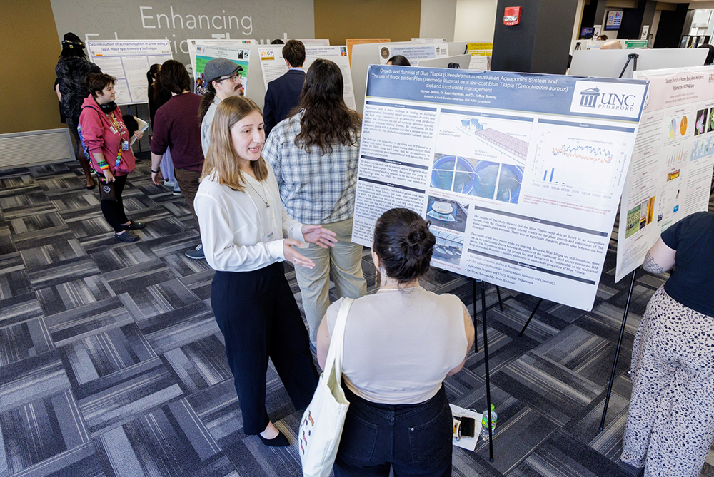 Overhead view of an undgraduate researcher presenting her work to a faculty member duing a poster session at the symposium at UNC Pembroke.