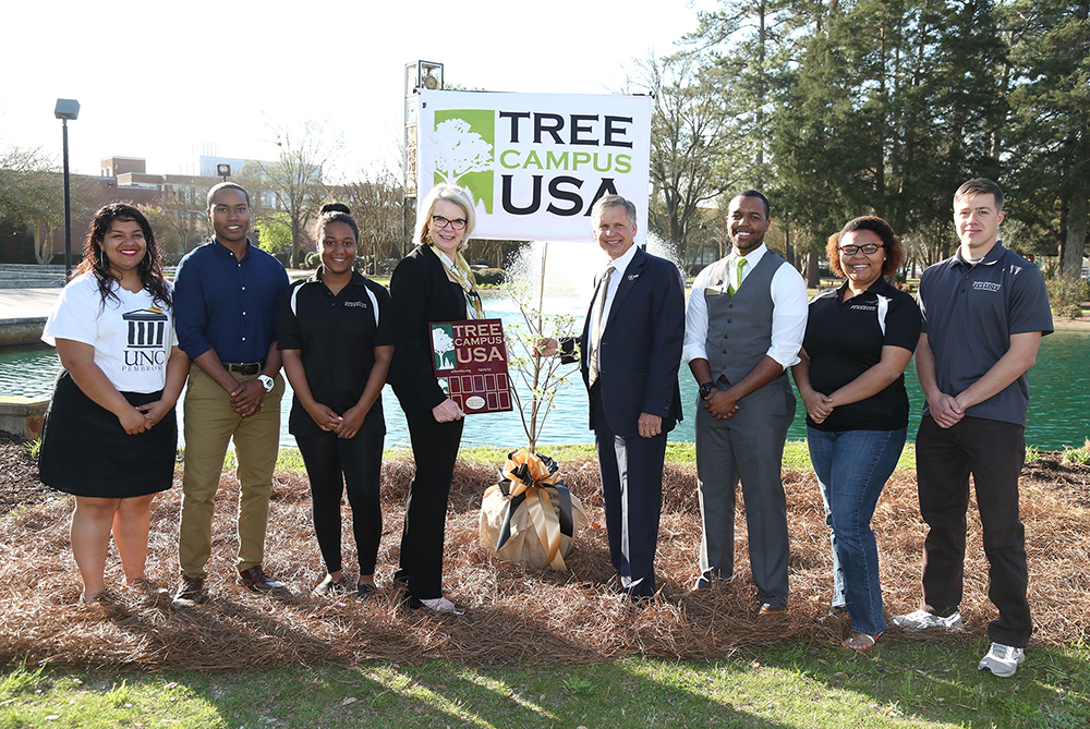 A group of eight people stands smiling in front of a “Tree Campus USA” banner on a university campus near a small pond. The individuals, including students and faculty, are gathered around a newly planted tree wrapped with a gold and black bow.