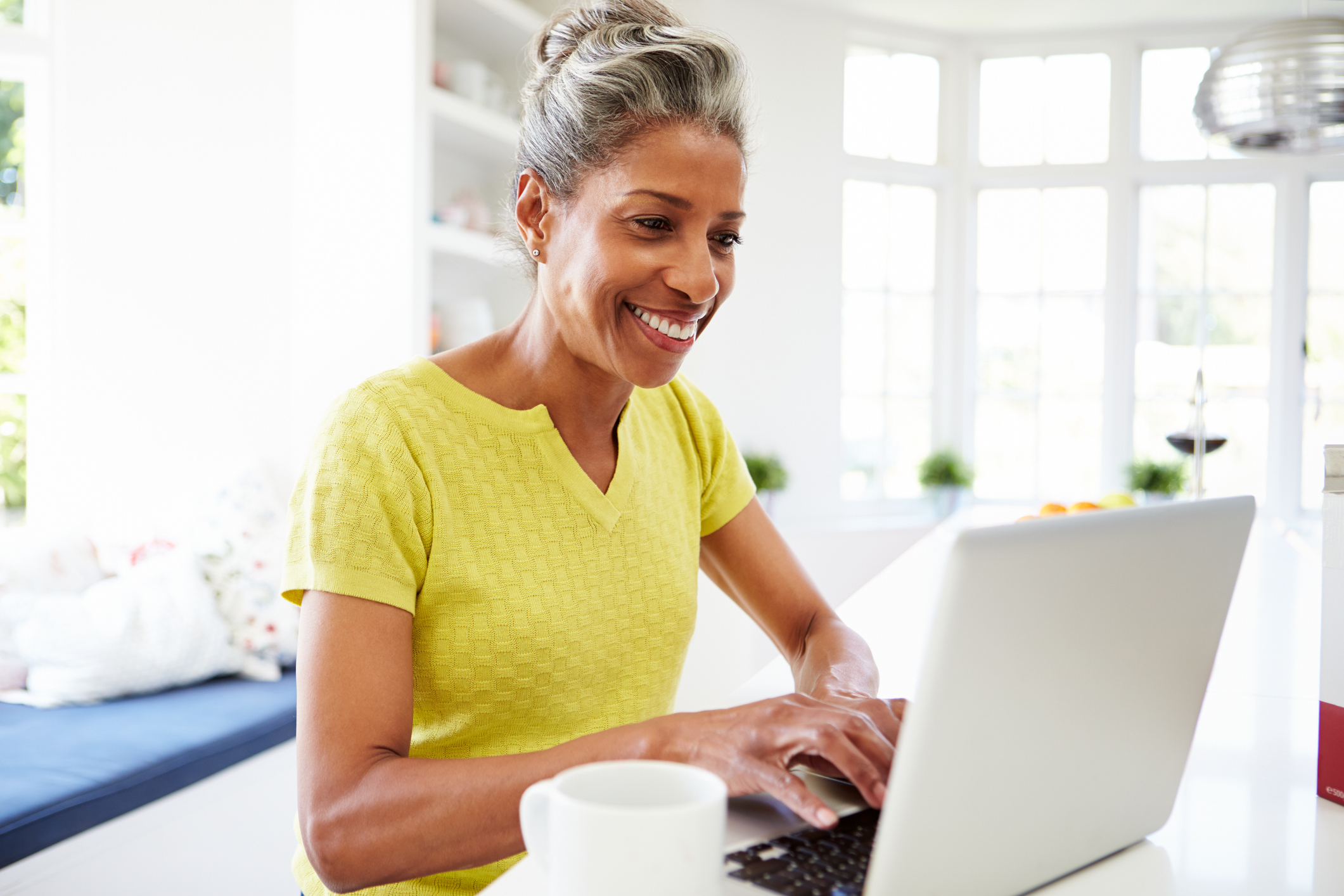 A person with grey hair wearing a yellow shirt, sitting at a table and typing on a laptop. A white coffee mug is in the foreground. The background includes large windows letting in natural light and shelves with various items. The person's face is obscured for privacy.
