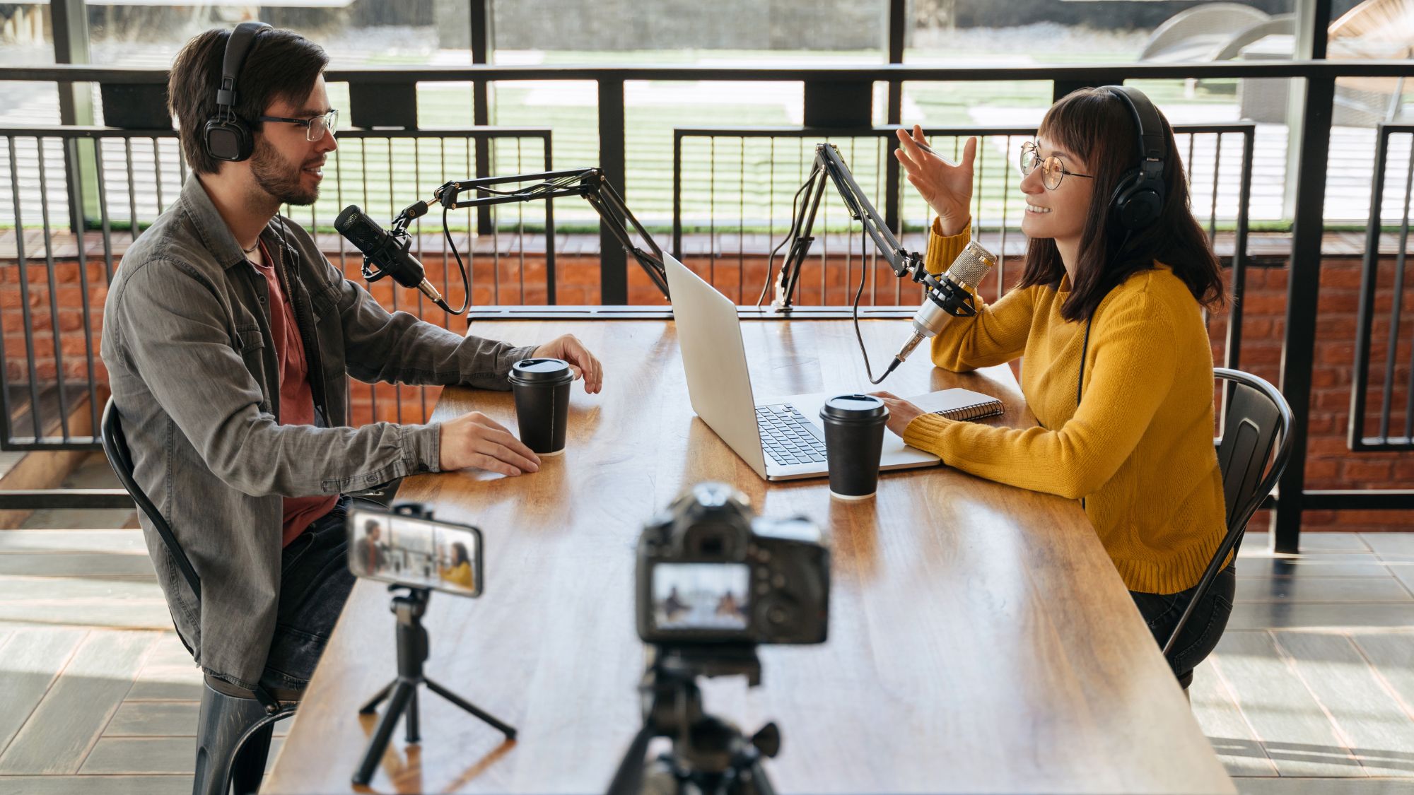 A man and woman sitting at a table recording a podcast with cameras and microphones.