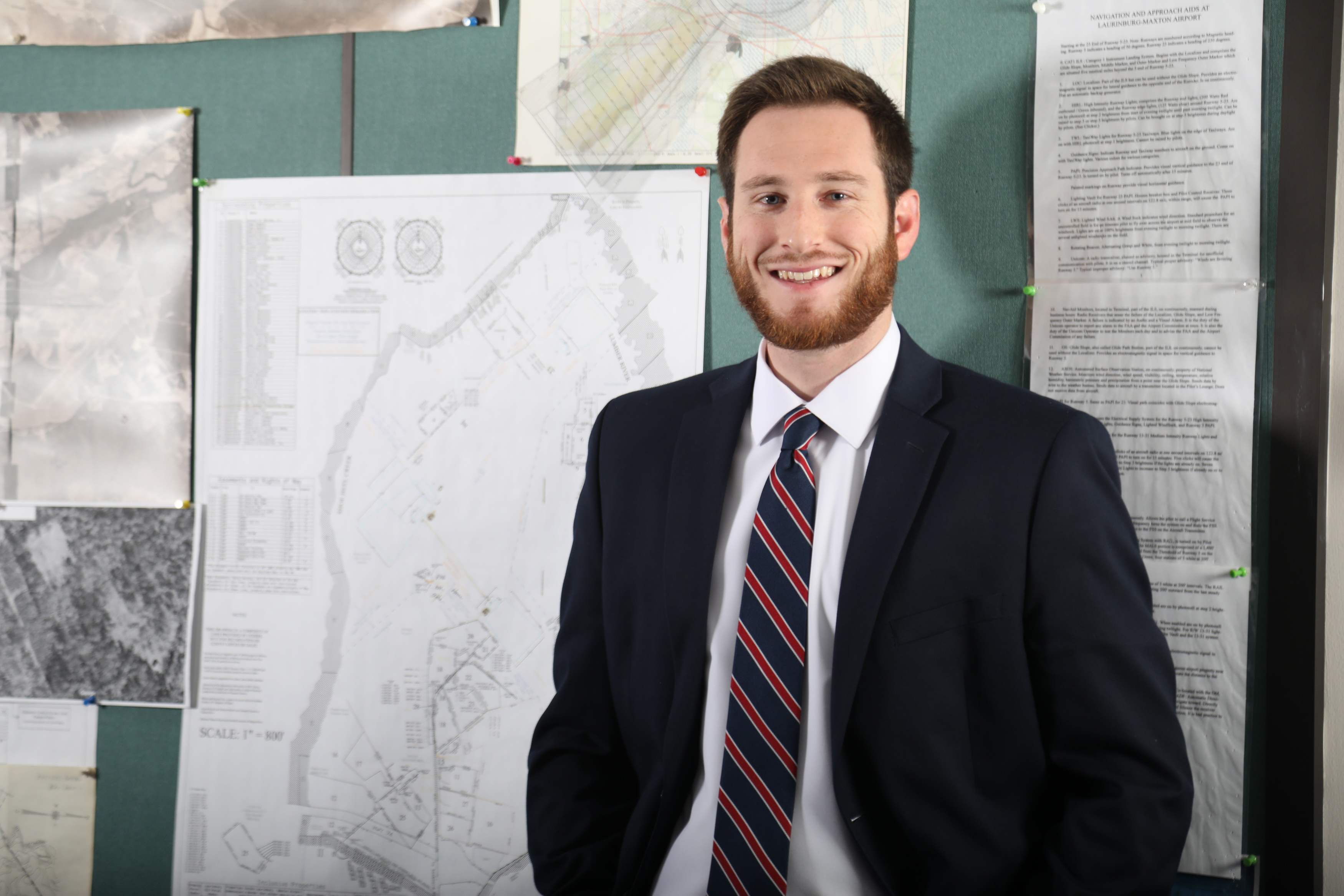 UNCP Business Administration alumnus Seth Hatchell stands in front of airport navigation charts at the Laurinburg-Maxton Airport, where he serves as Executive Director, representing career outcomes for Business Administration graduates.