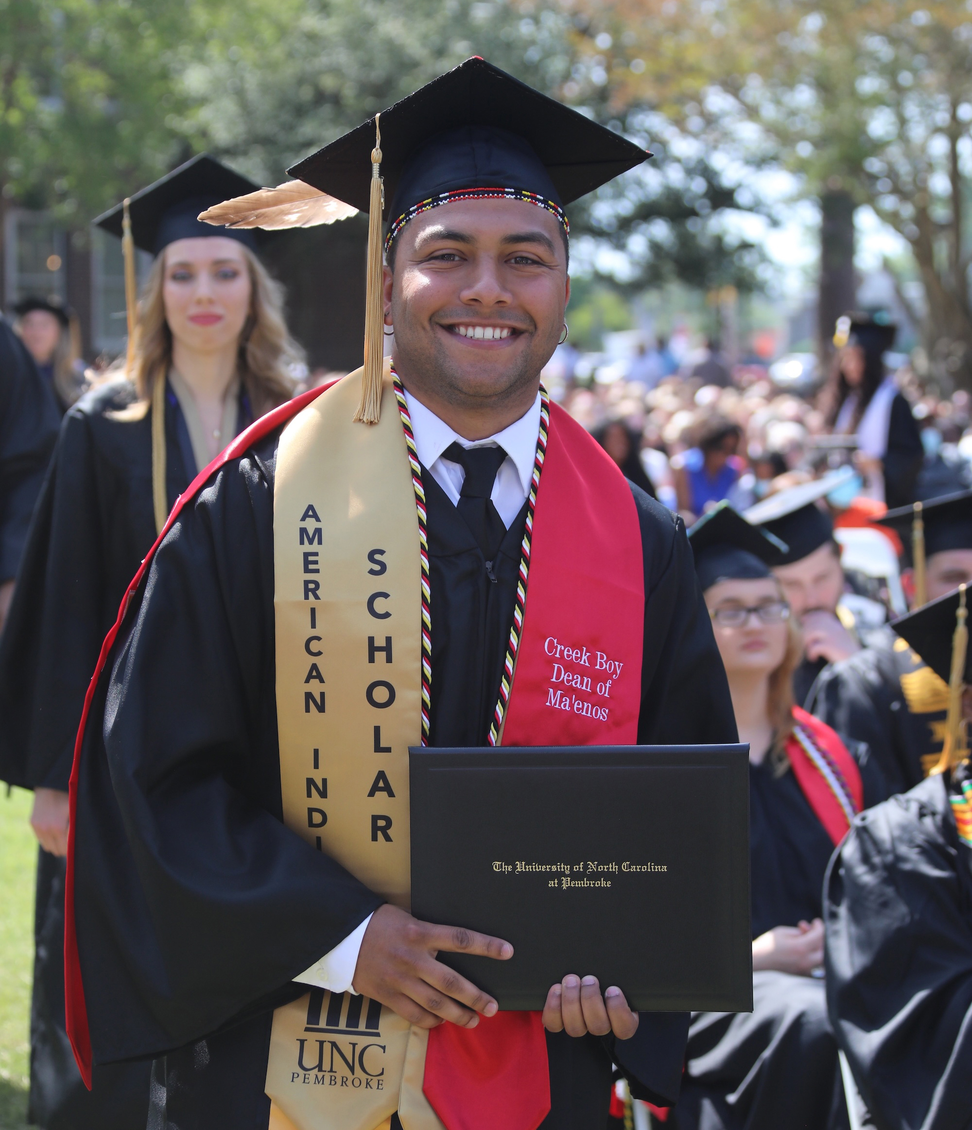 UNCP Criminal Justice graduate Cameron Richardson smiles at commencement holding his UNC Pembroke diploma, wearing an American Indian Scholar stole, a beaded cap band, and a red stole reading "Creek Boy Dean of Ma'enos," representing graduate success in the program.