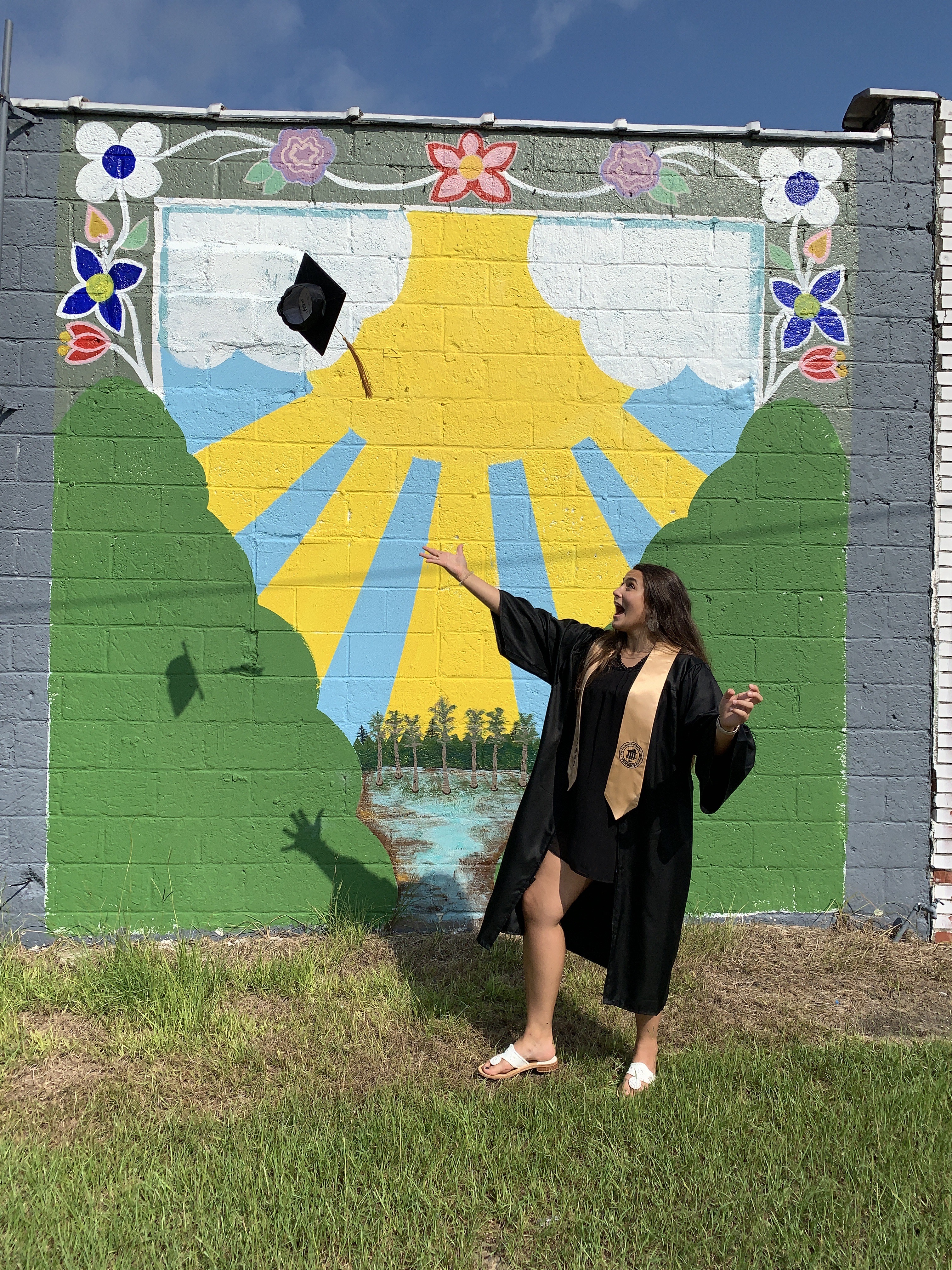 Female student in her cap and gown in front of a mural she painted on a building downtown Pembroke. The mural shows trees, the sun and water.