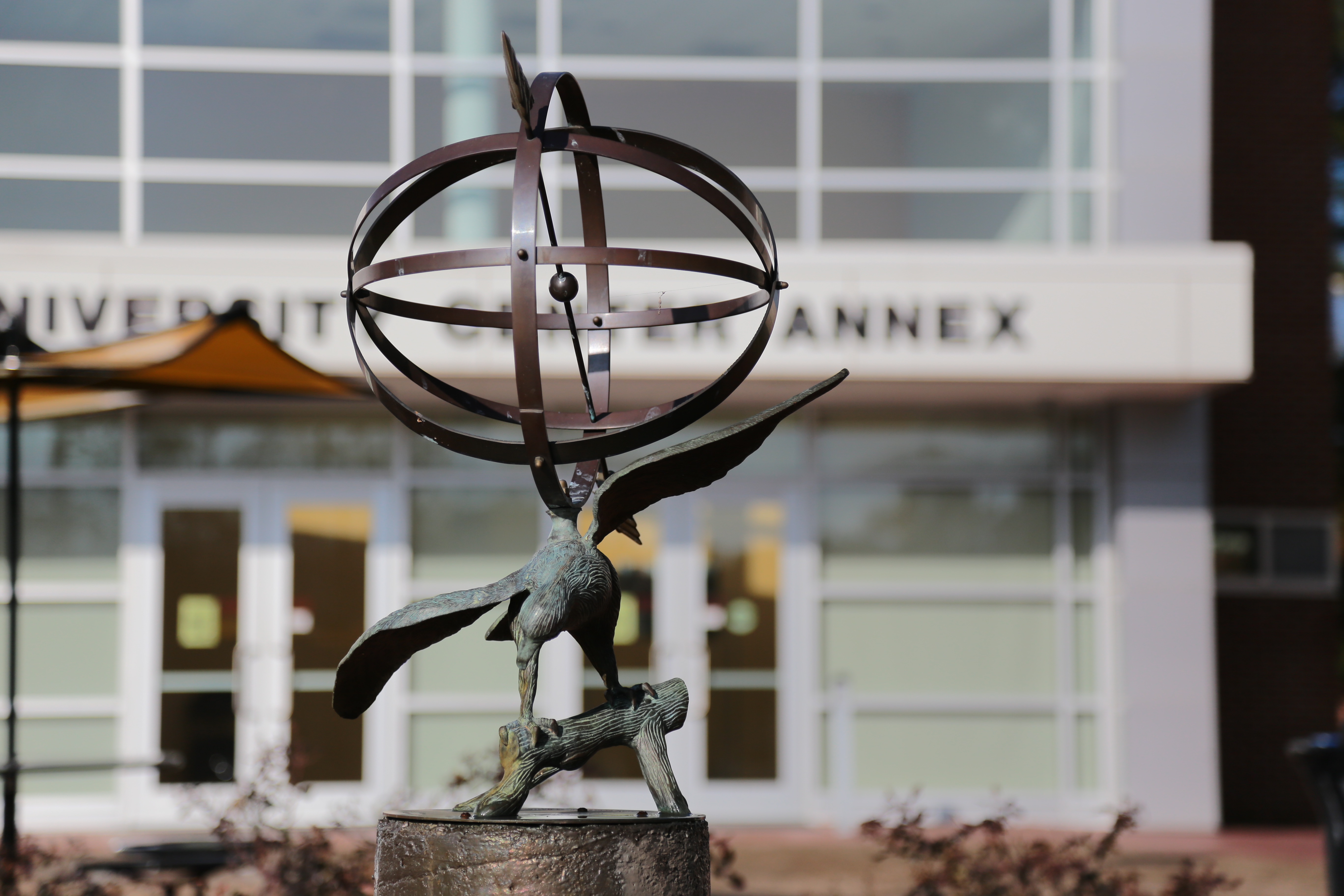 A sculpture of an eagle with outstretched wings perched on a branch, supporting an armillary sphere. In the background, a building with large windows and the partially visible text 'UNIVERSITY ANNEX' can be seen.
