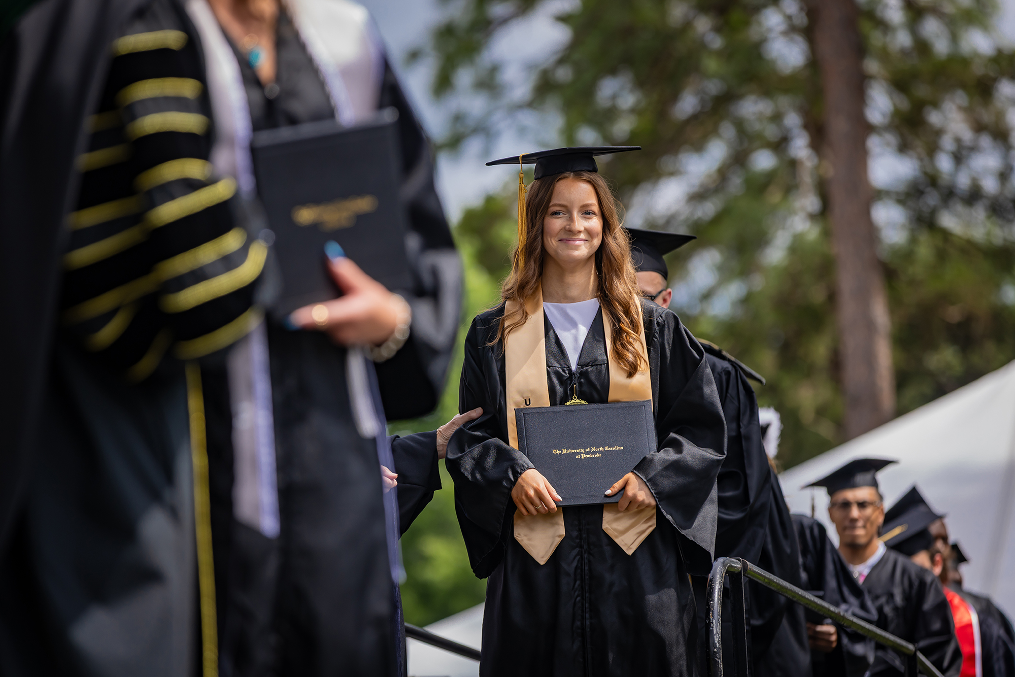 A 2026 UNCP graduate in cap and gown holds her diploma and leads a line of graduates across the commencement stage.