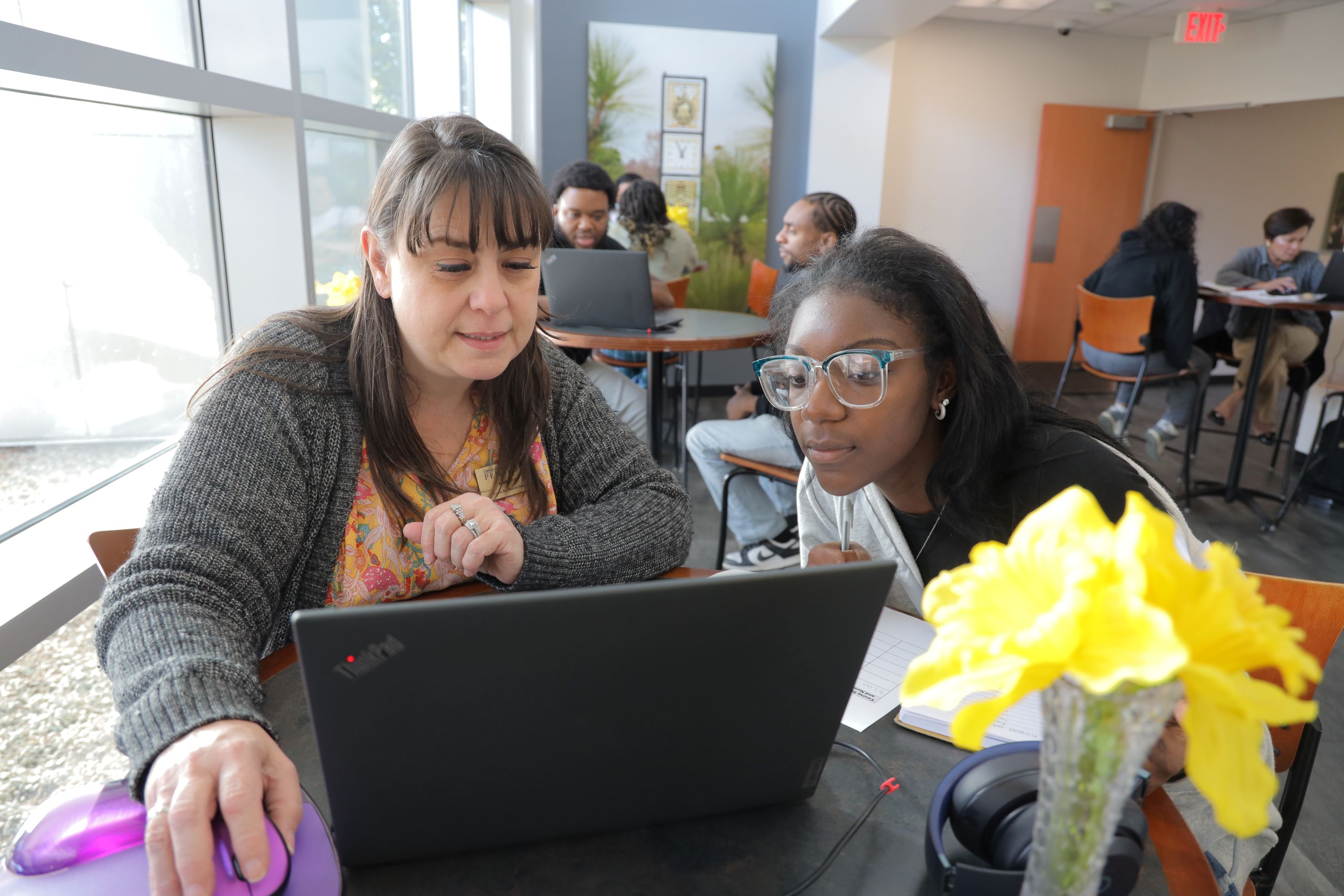 Female advisor and student, together looking at a laptop on a bistro table with other advisors and students similarly seated around the room for advising day at UNC Pembroke