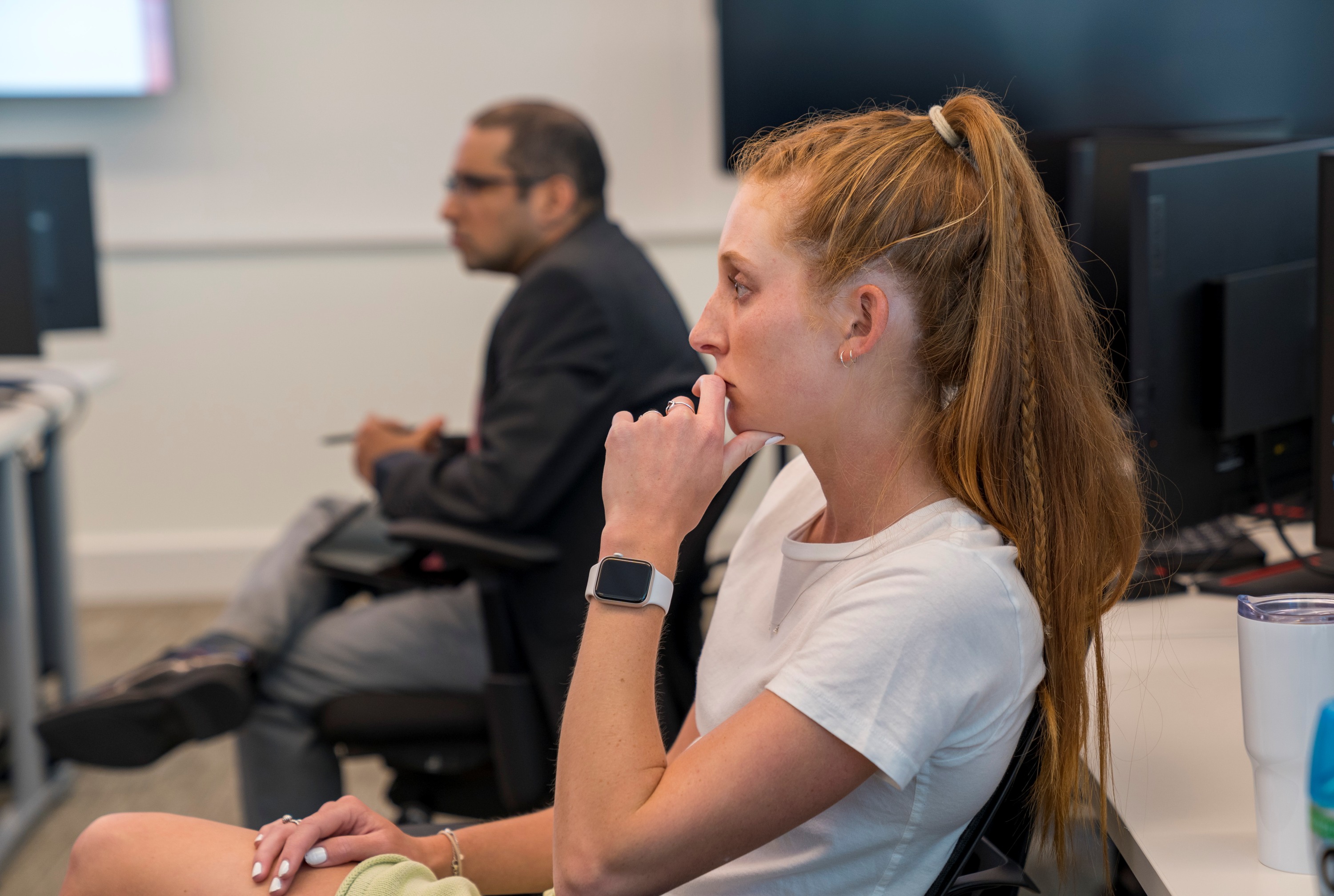 Female student in a classroom, attentively listening to a student presentation with her chin resting on her hand. The professor is visible, softly blurred, in another seat to her side.