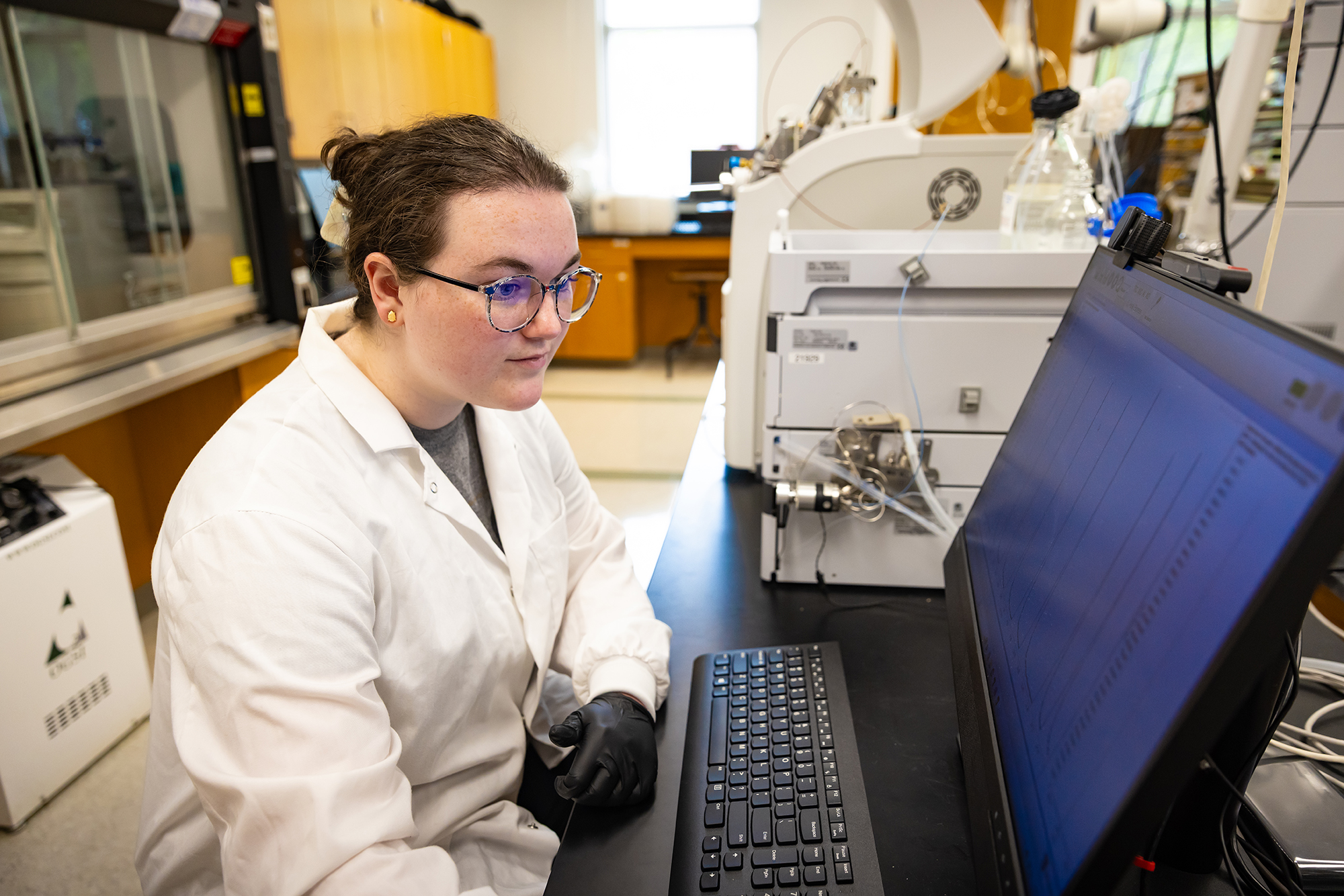 female student in labcoat at UNCP sitting in a lab in front of a computer