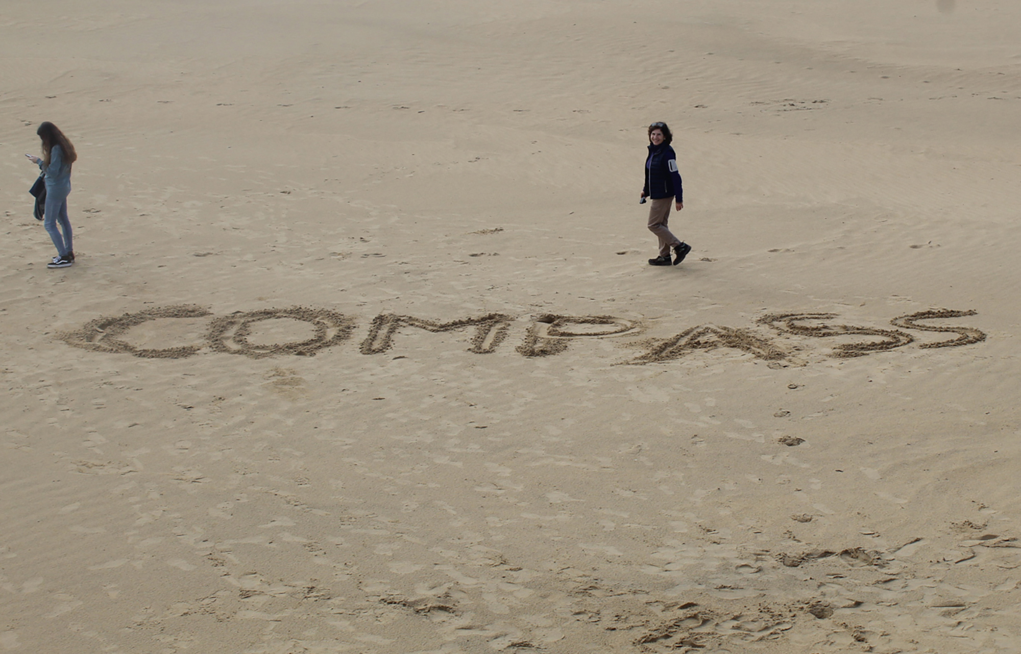 three students walk on a beach around the word "COMPASS" drawn in the sand